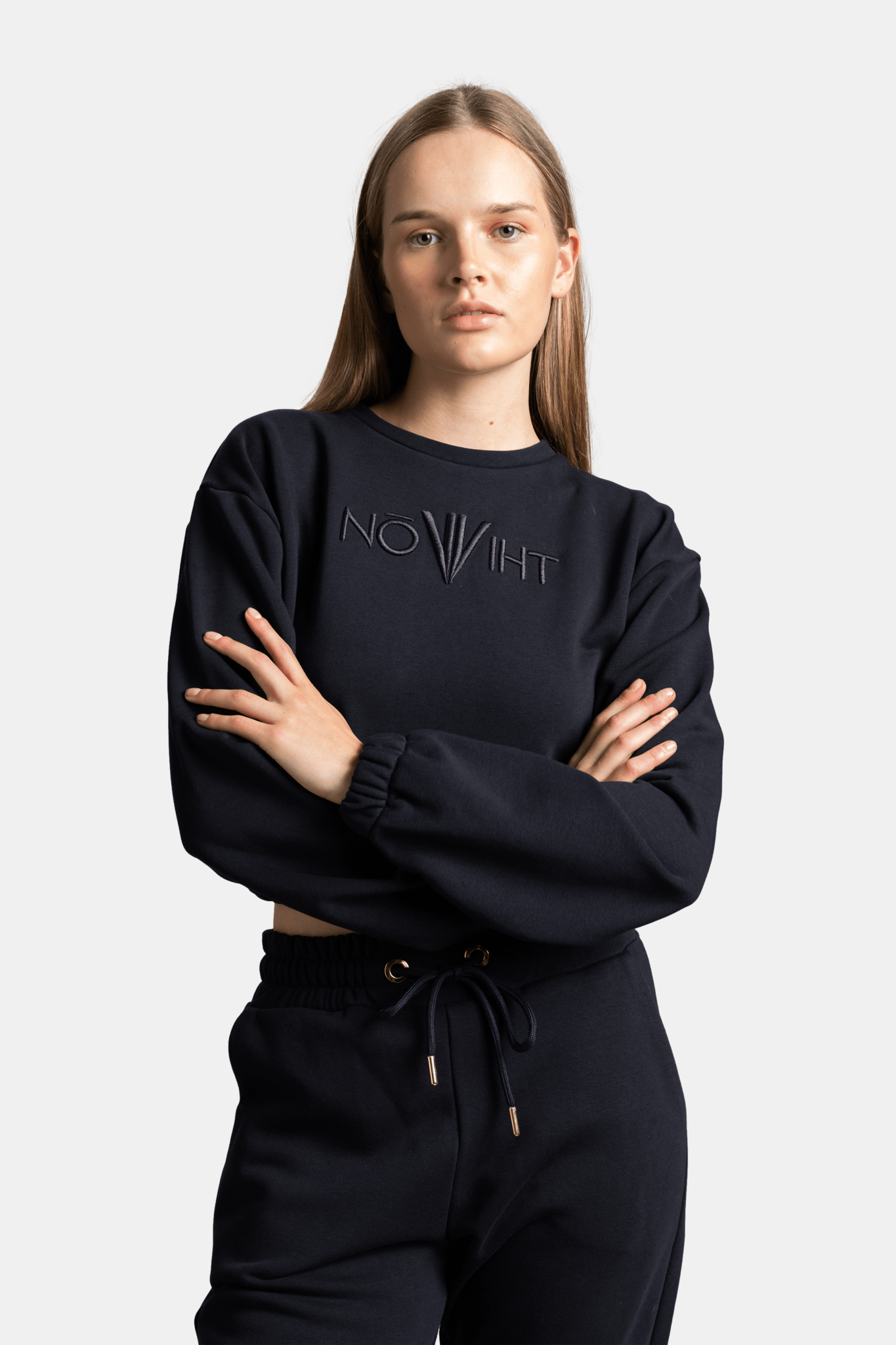 Close-up of the woman wearing a black tracksuit top with embossed logo, crossing her arms, focusing on the chest and logo detail.