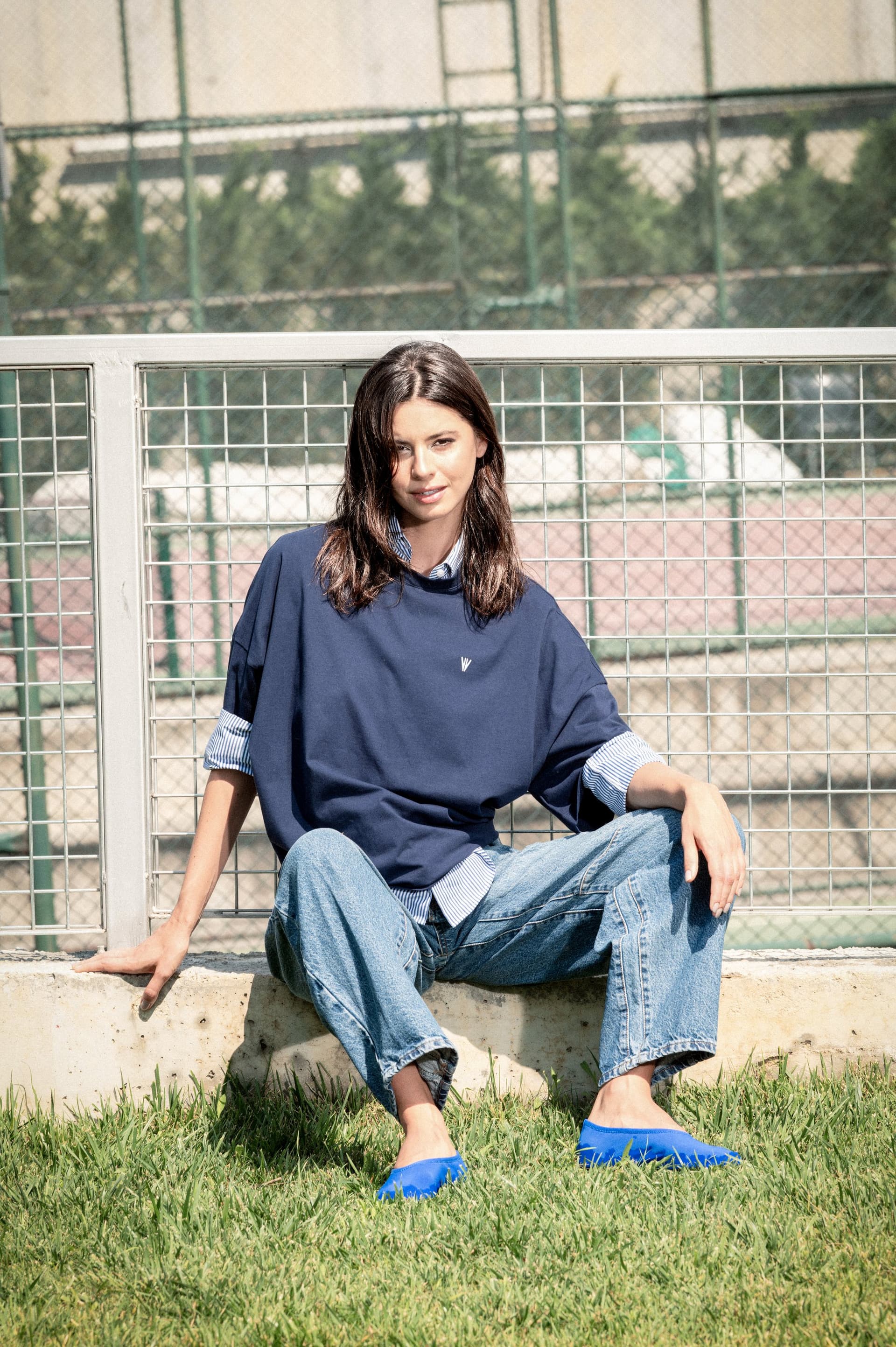 Model in a navy NOWIHT sweatshirt over a light blue shirt, sitting by a tennis court