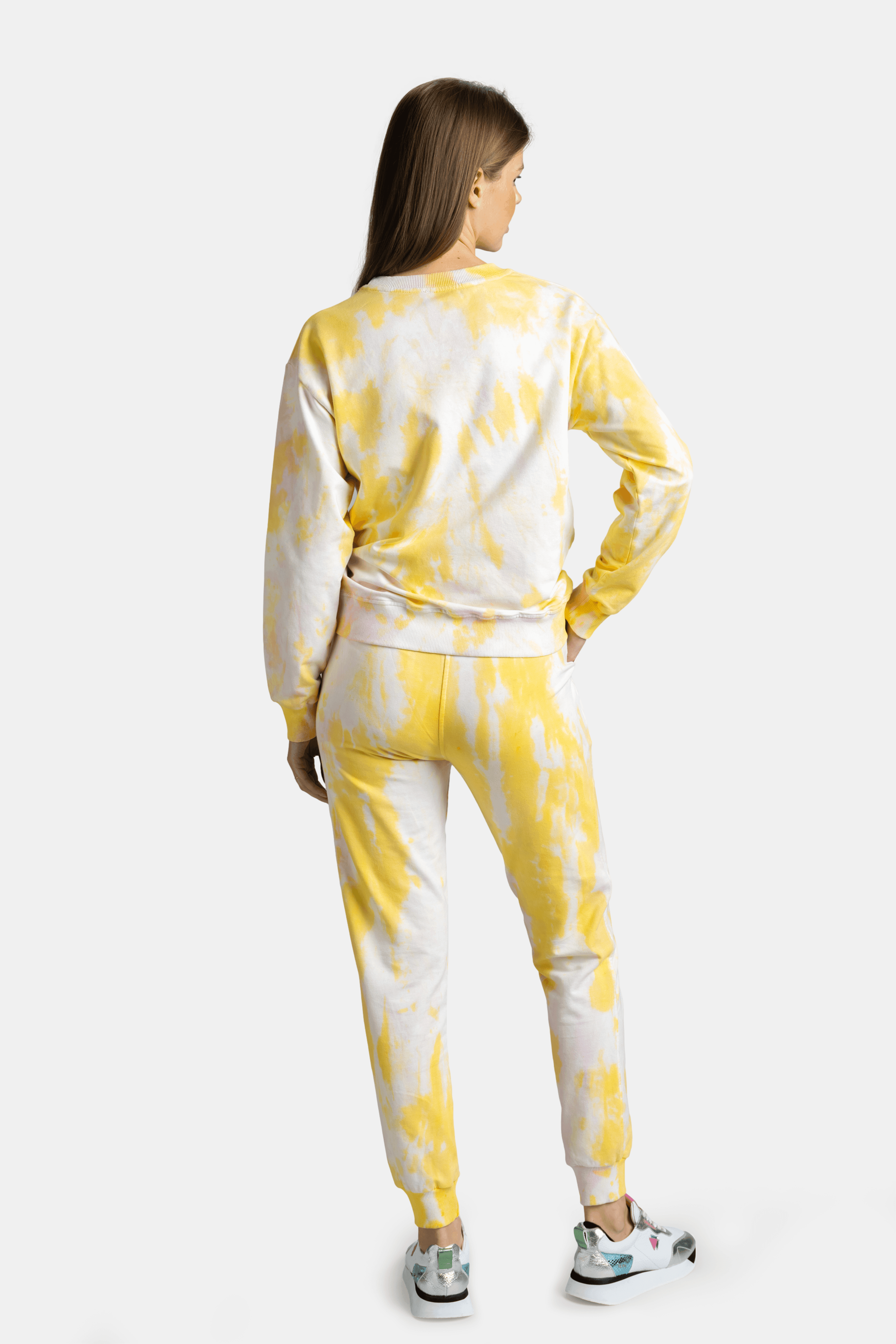 Side angle of a woman in a yellow tie-dye tracksuit, hands in pockets, looking slightly over her shoulder.