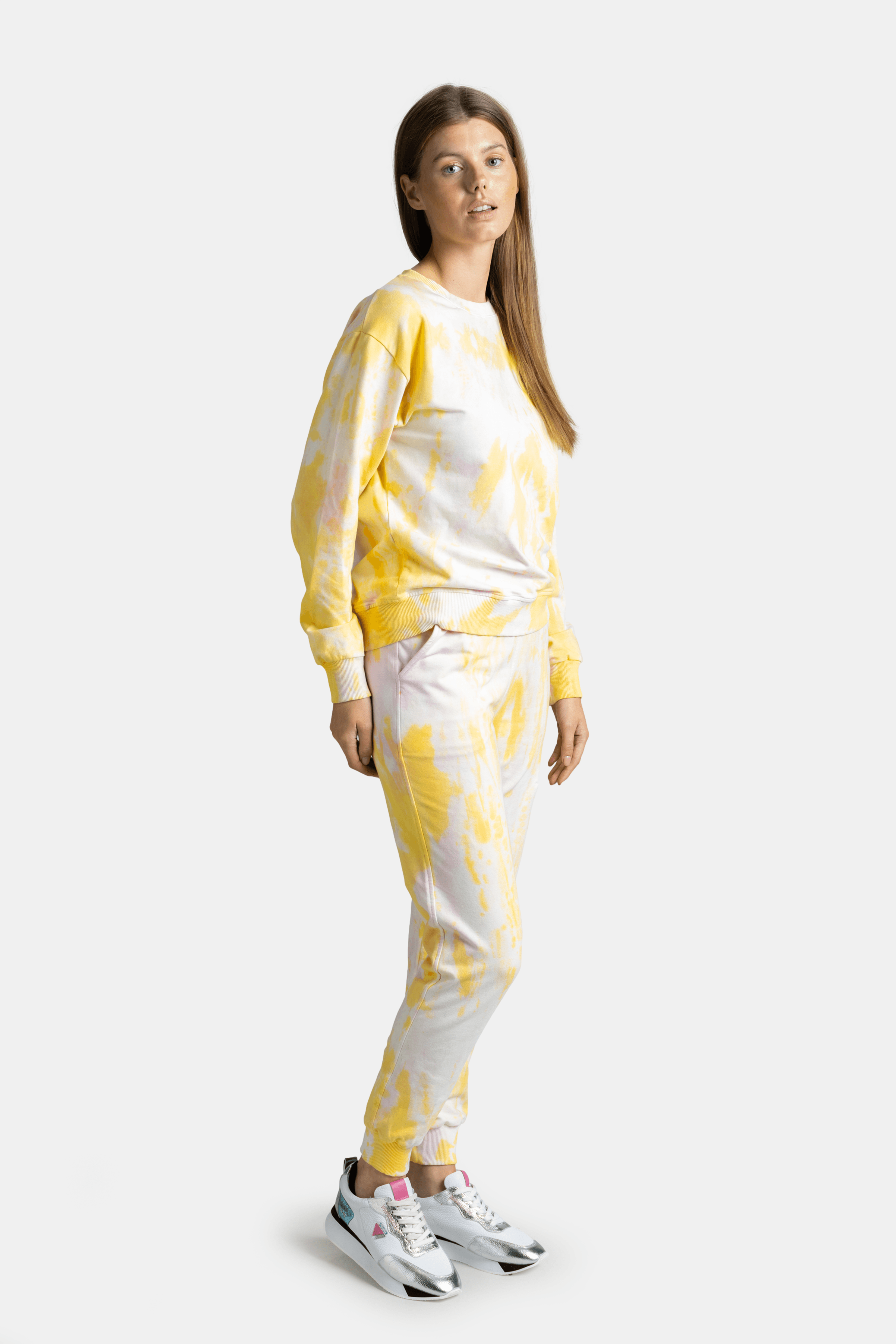 Woman in a yellow tie-dye tracksuit, side profile with hands in pockets, looking ahead.