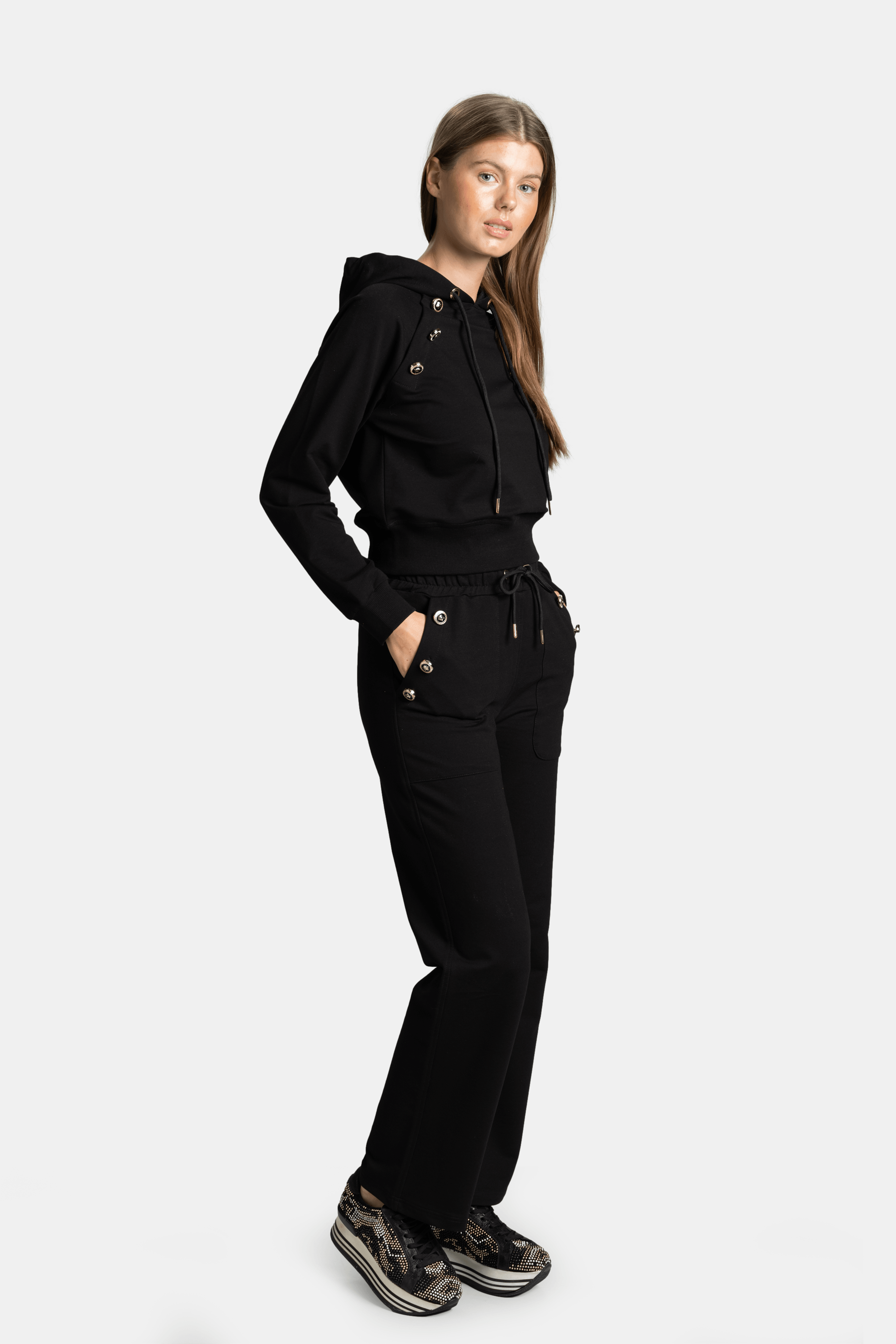 Side profile of a woman in a black hoodie and pants set with decorative gold accents, hands in pockets.