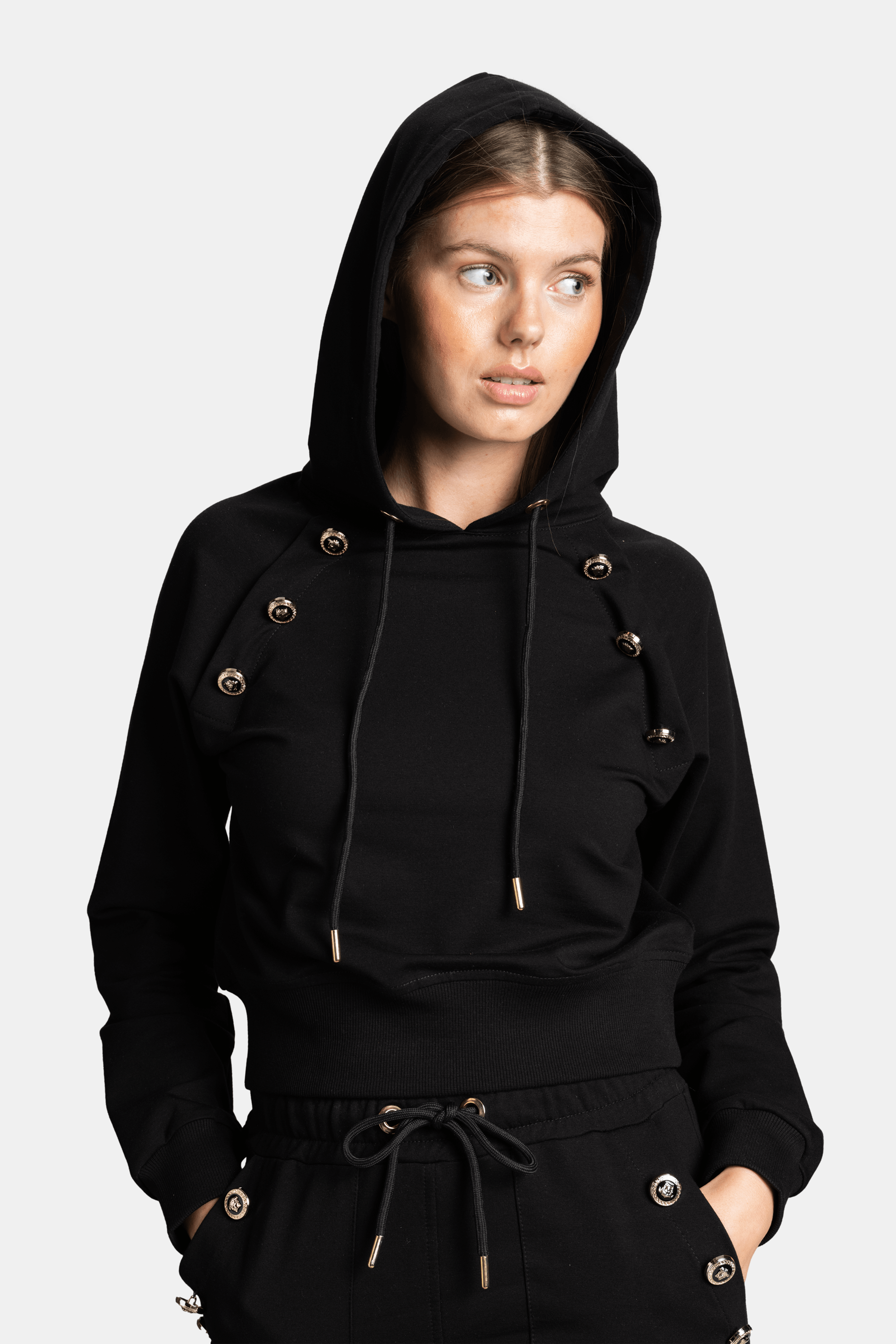 Close-up of a woman in a black hoodie with decorative gold buttons, hood up, looking to the side.