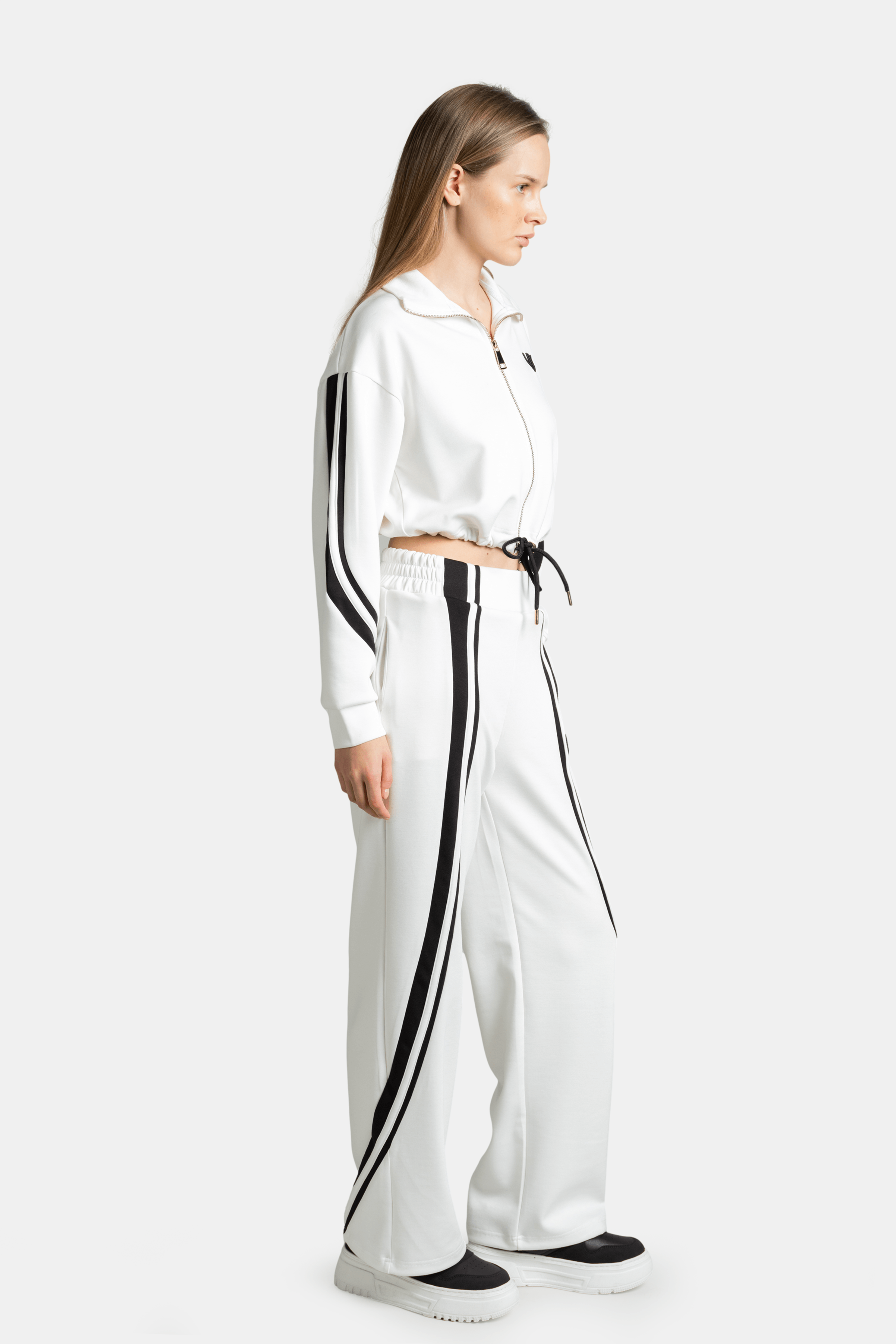 Side view of a woman in a white tracksuit set with black side stripes, highlighting the relaxed fit and long pants.