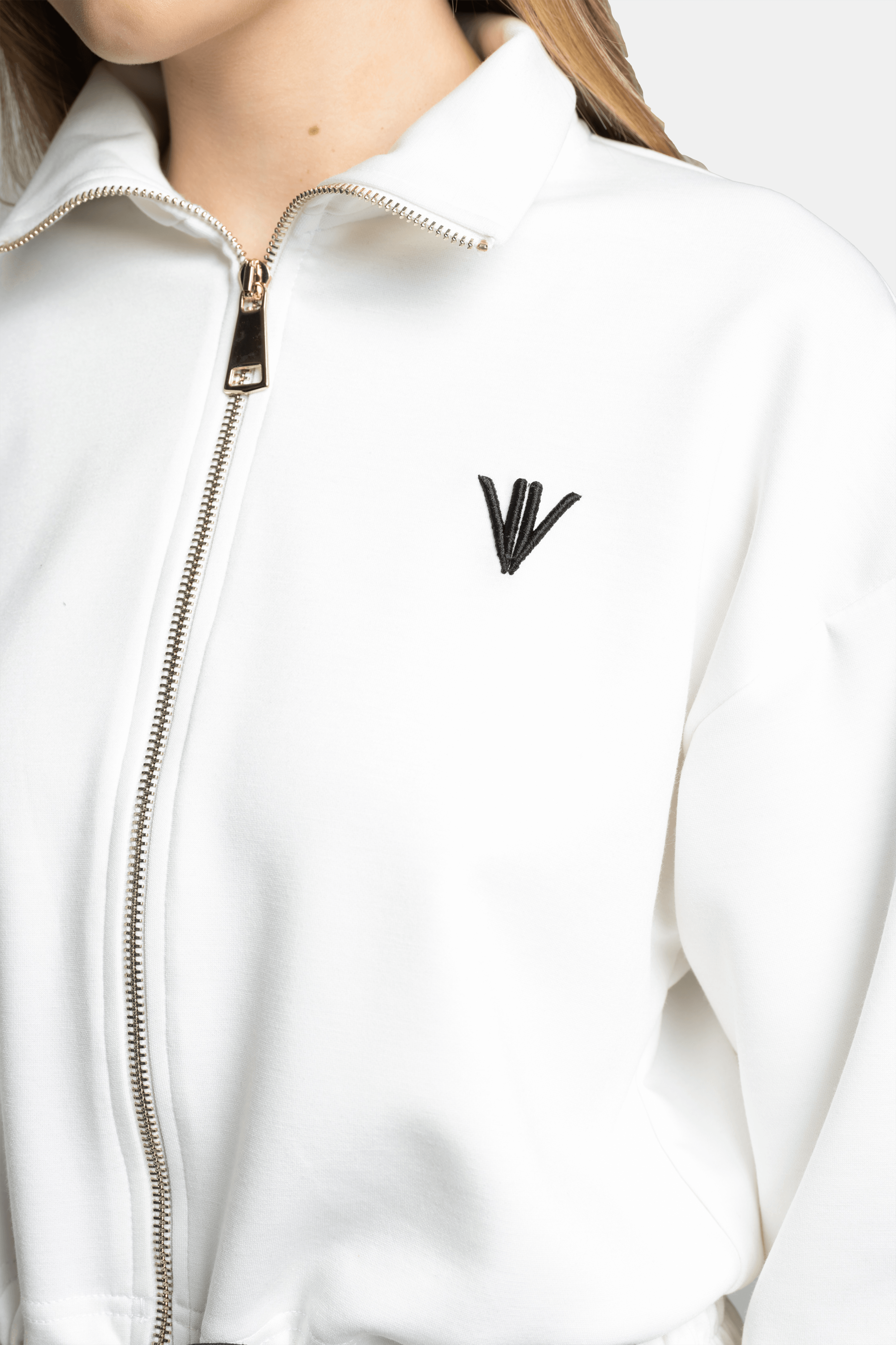 Close-up of the embroidered logo on the chest of the white zip-up tracksuit top with black accents.