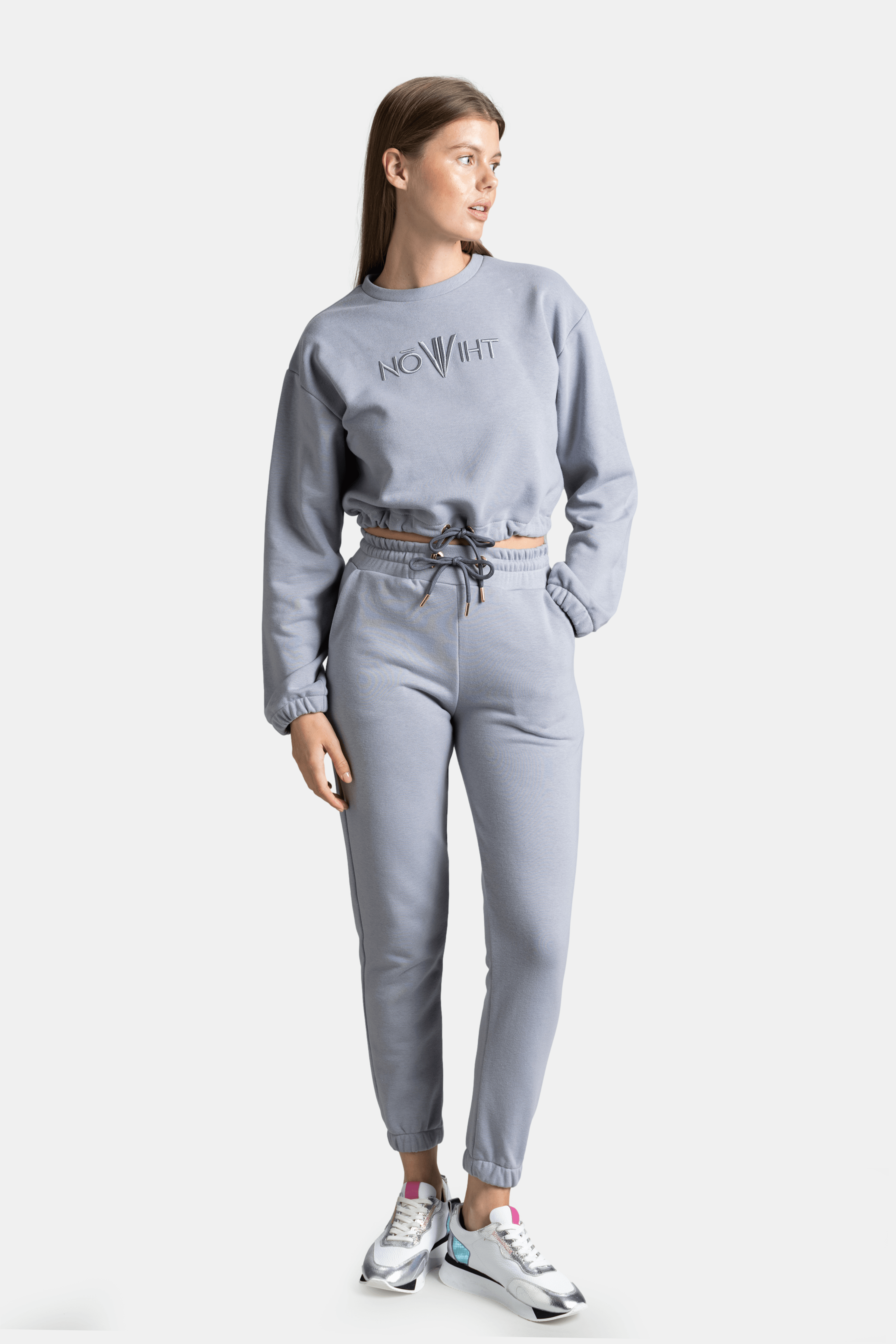 Woman in light gray NOWIHT tracksuit set, side view, looking to the side, hands in pockets.