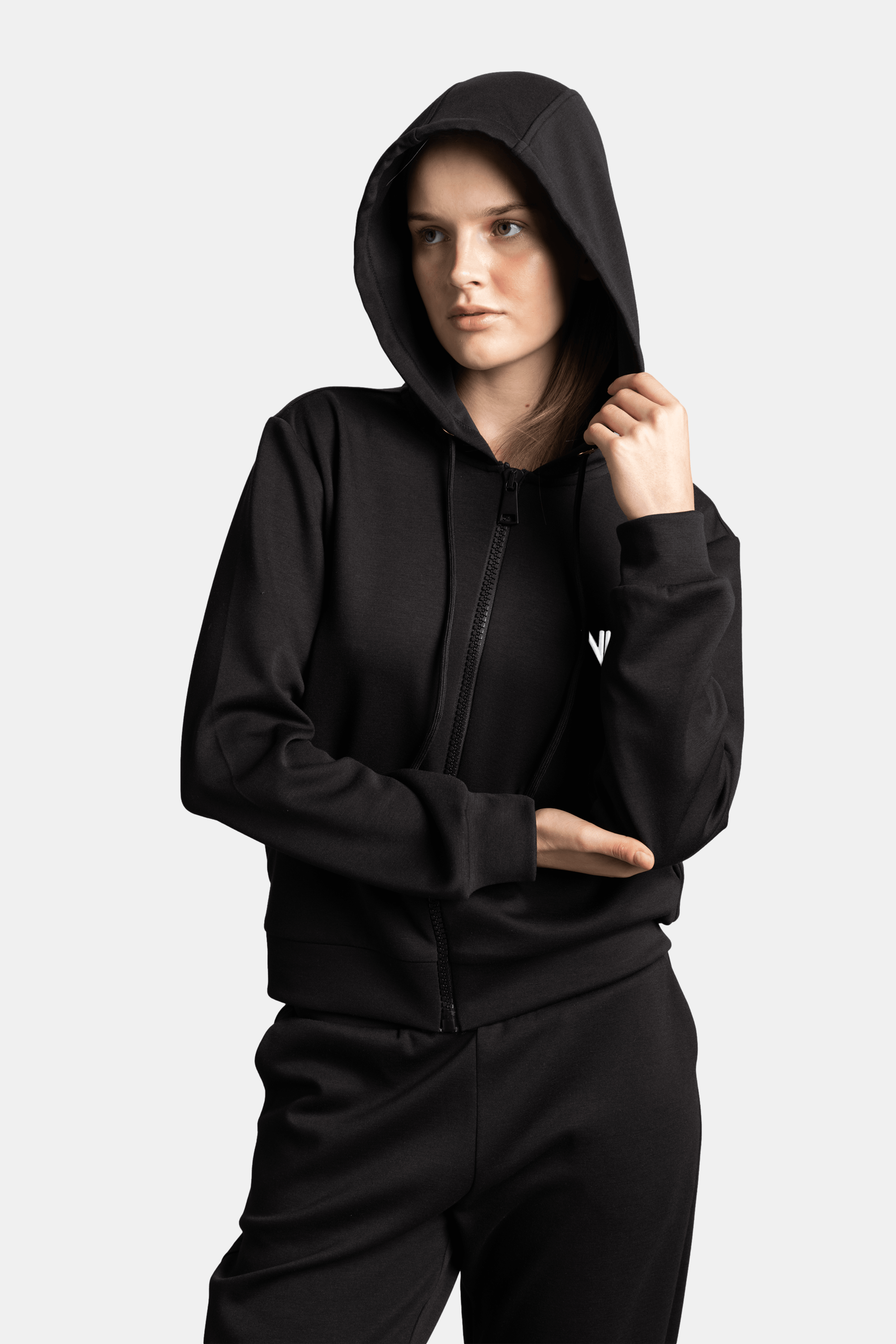 Woman in a black hoodie with the hood up, crossing her arms, with a confident expression and the hoodie’s front zipper in view.