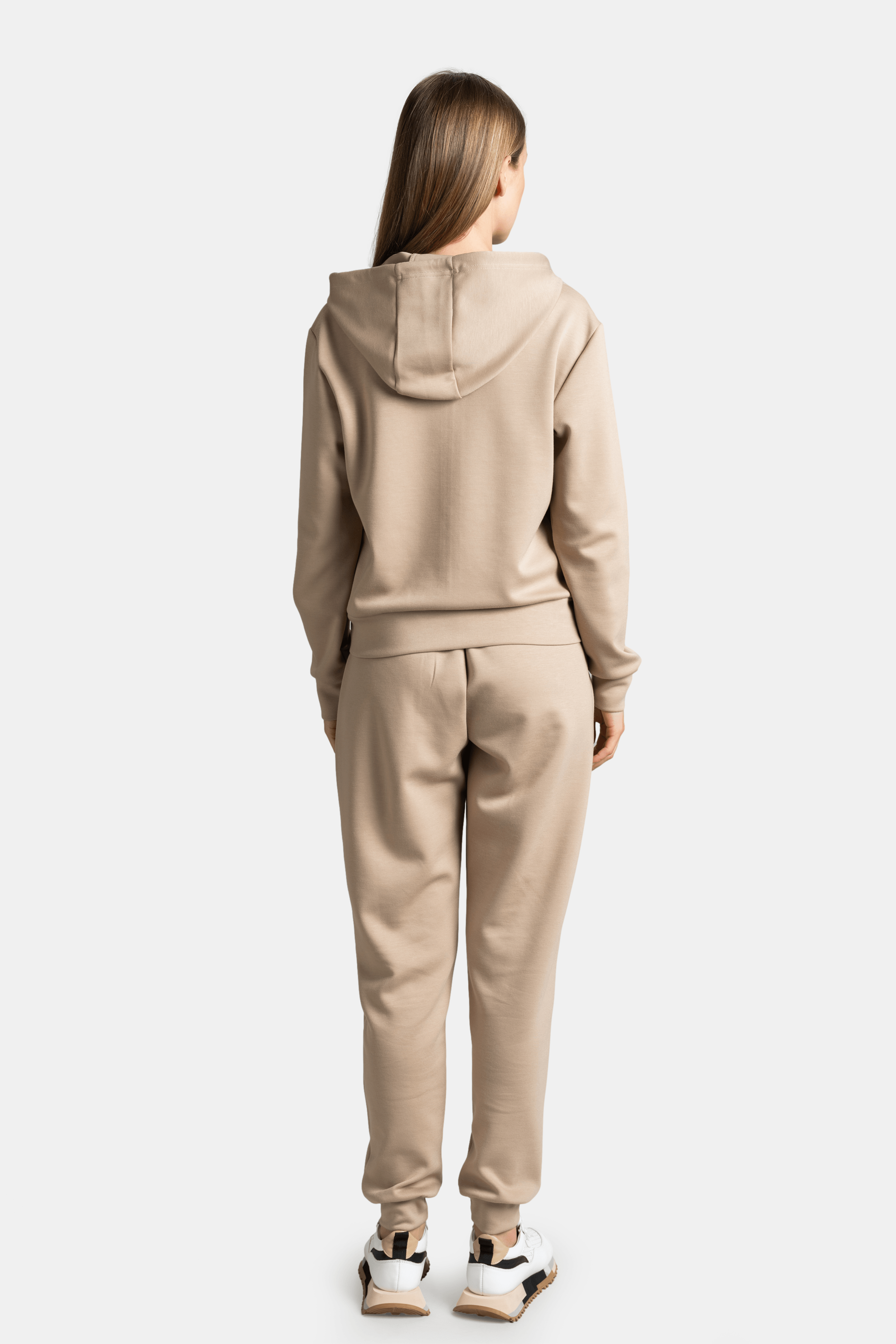 Back view of a woman wearing a beige hoodie tracksuit, showing the hood and relaxed fit of the outfit.