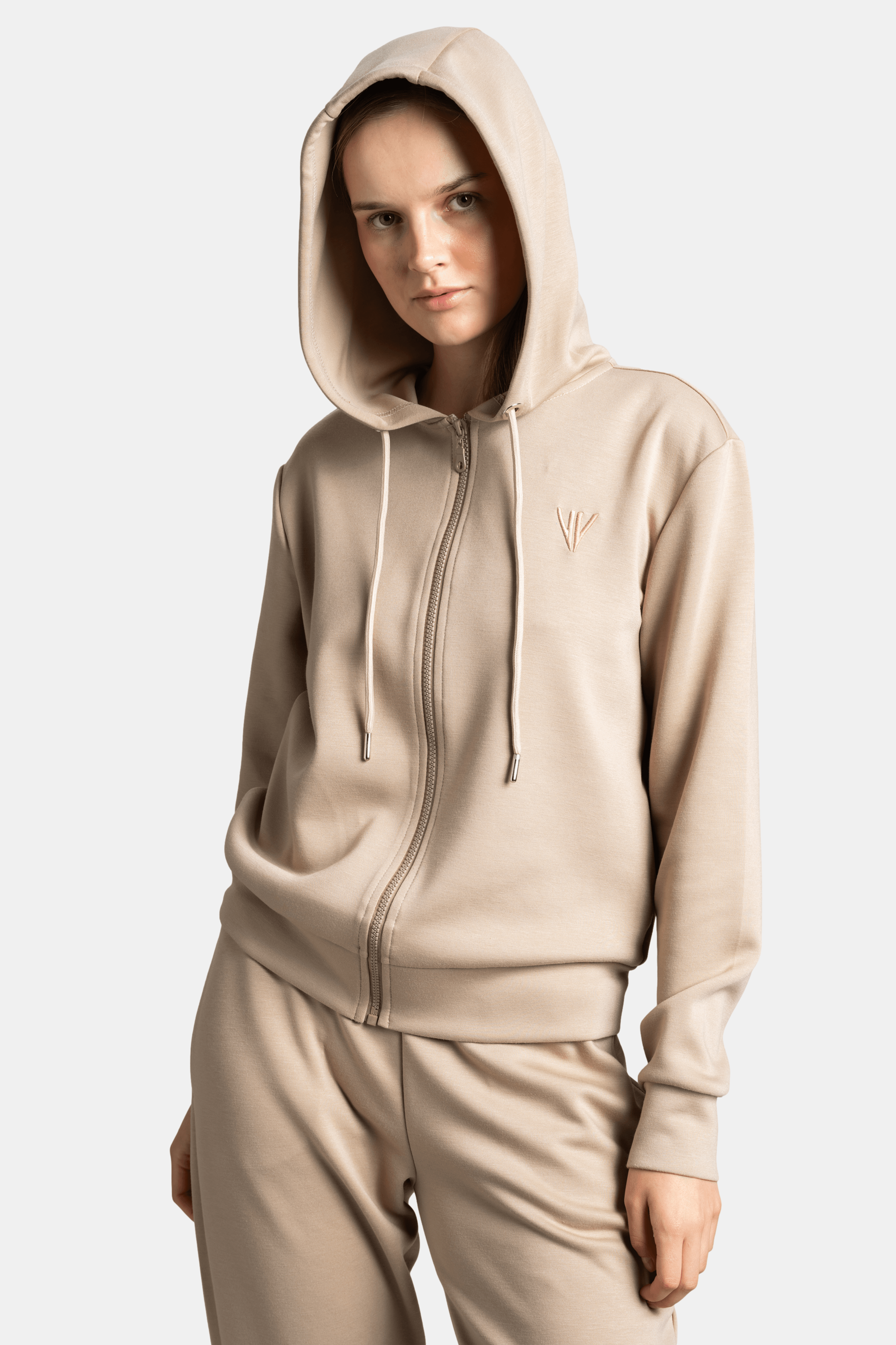 Close-up of a woman in a beige hoodie tracksuit, with the hood up, looking slightly downward.