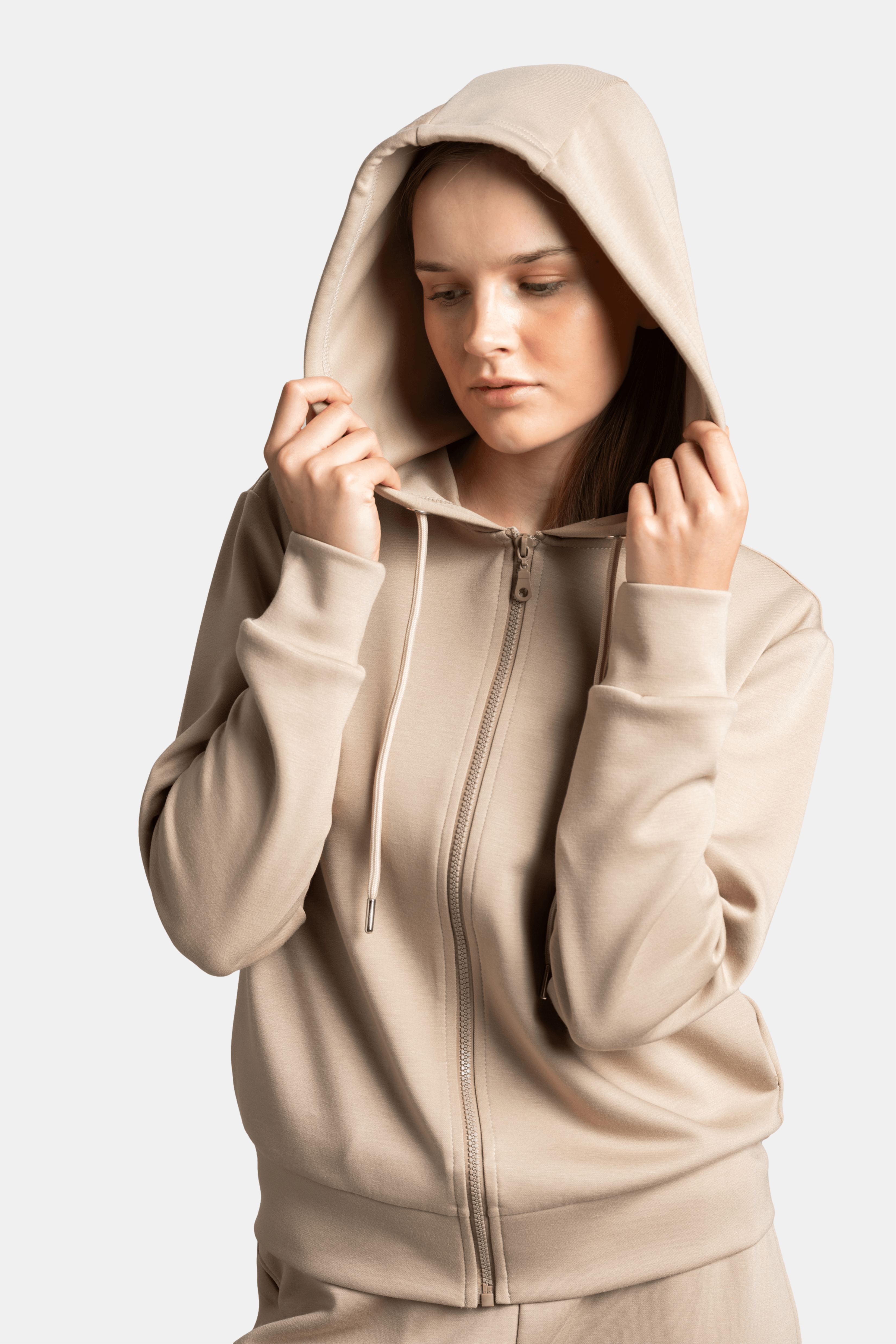 Woman gently holding the edges of her hood, wearing a beige hoodie tracksuit with a soft expression.