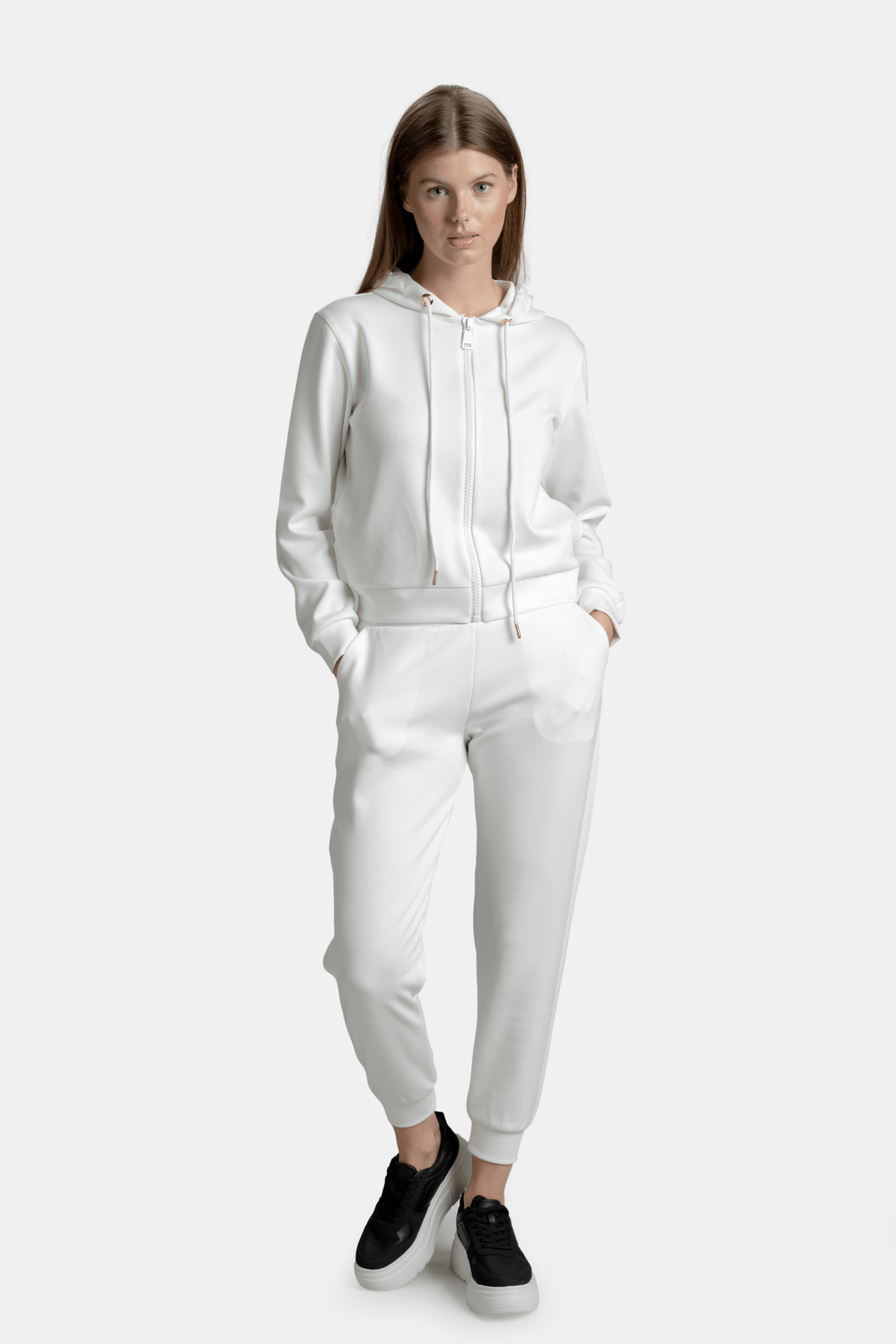 Front view of a woman wearing an elegant white tracksuit with a hood, hands in pockets, standing confidently.