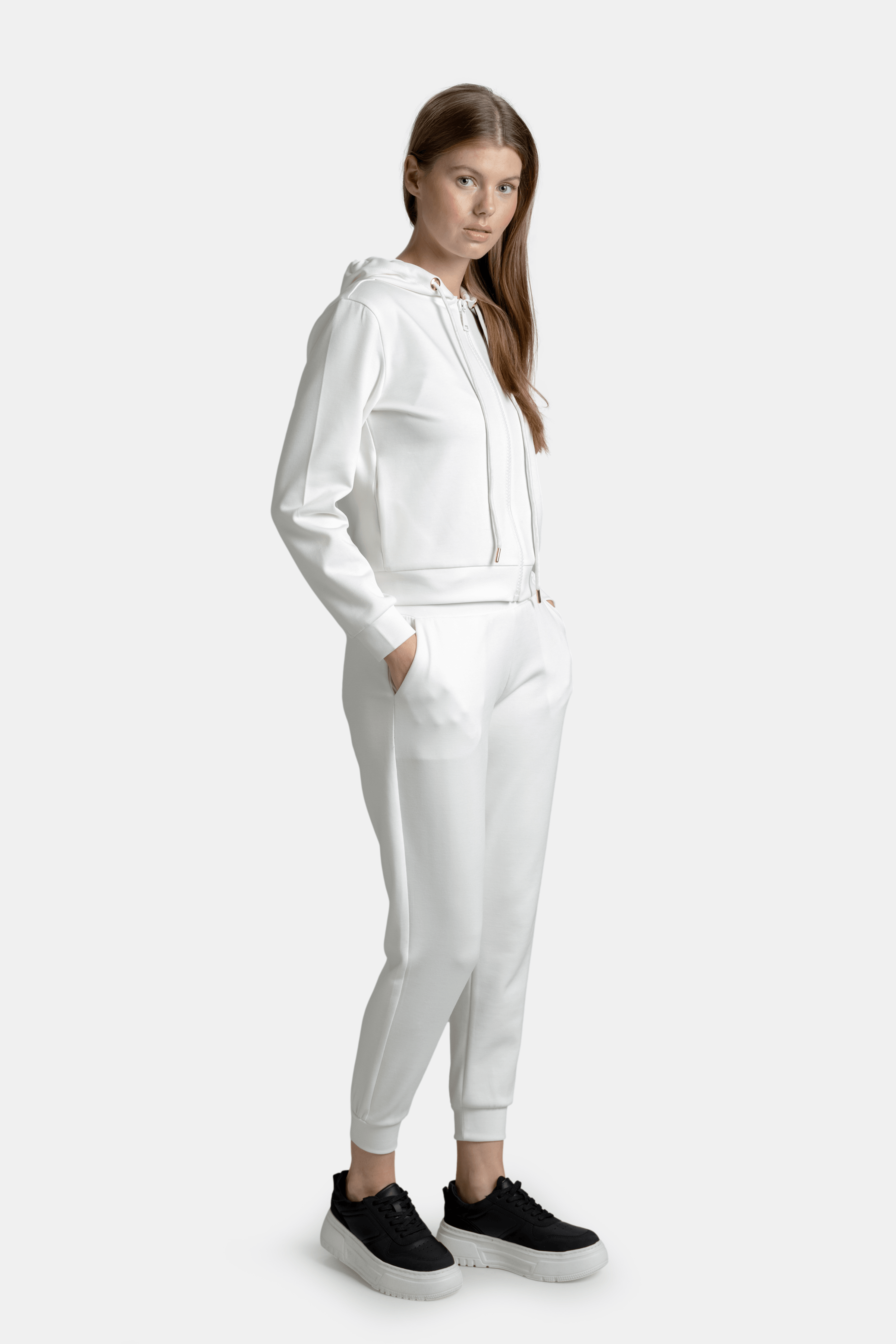 Side profile of a woman in a white hooded tracksuit, hands in pockets, standing in a relaxed pose.