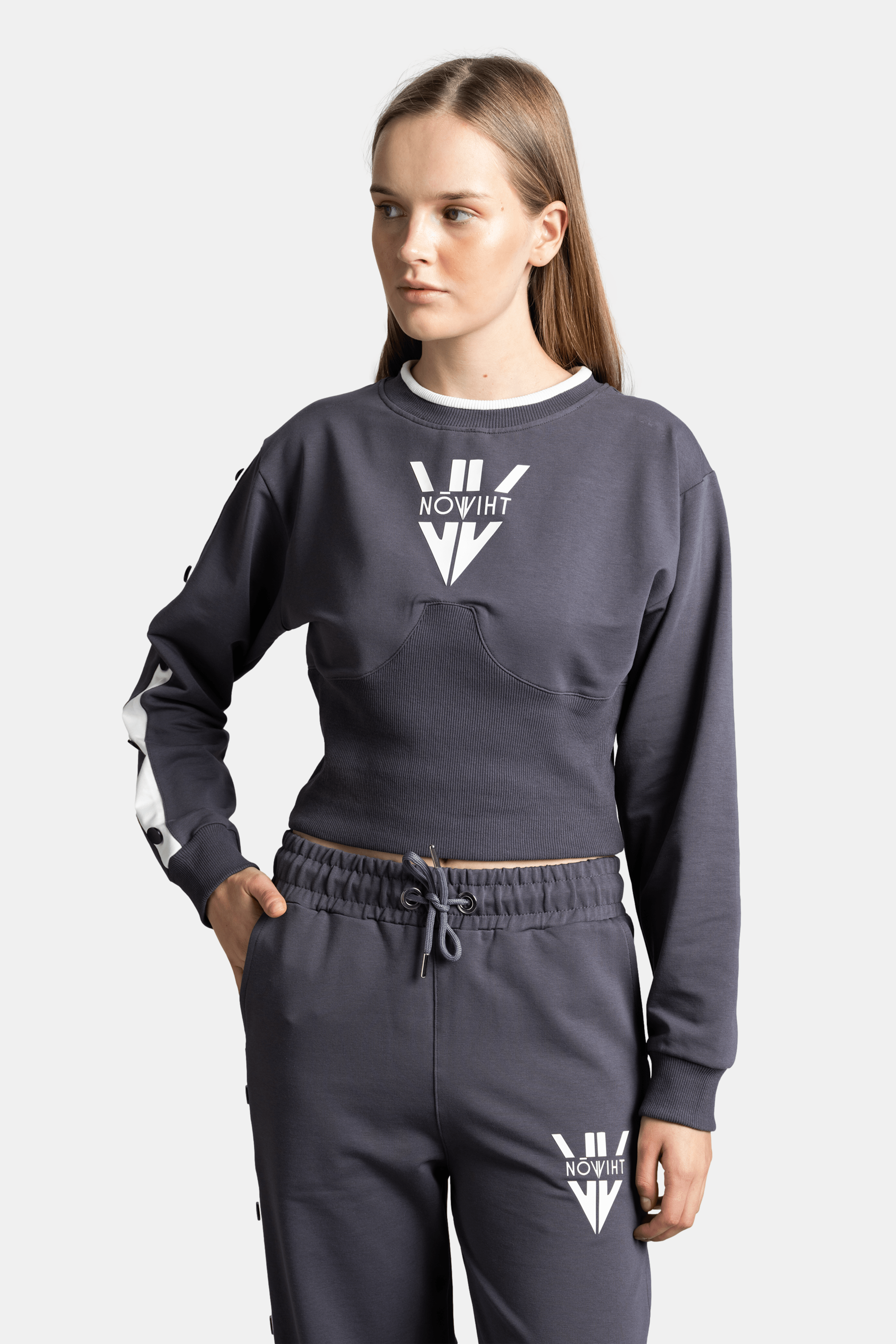 Woman in dark gray loungewear set, wearing a cropped sweatshirt and matching pants with white logo and stripe accents.