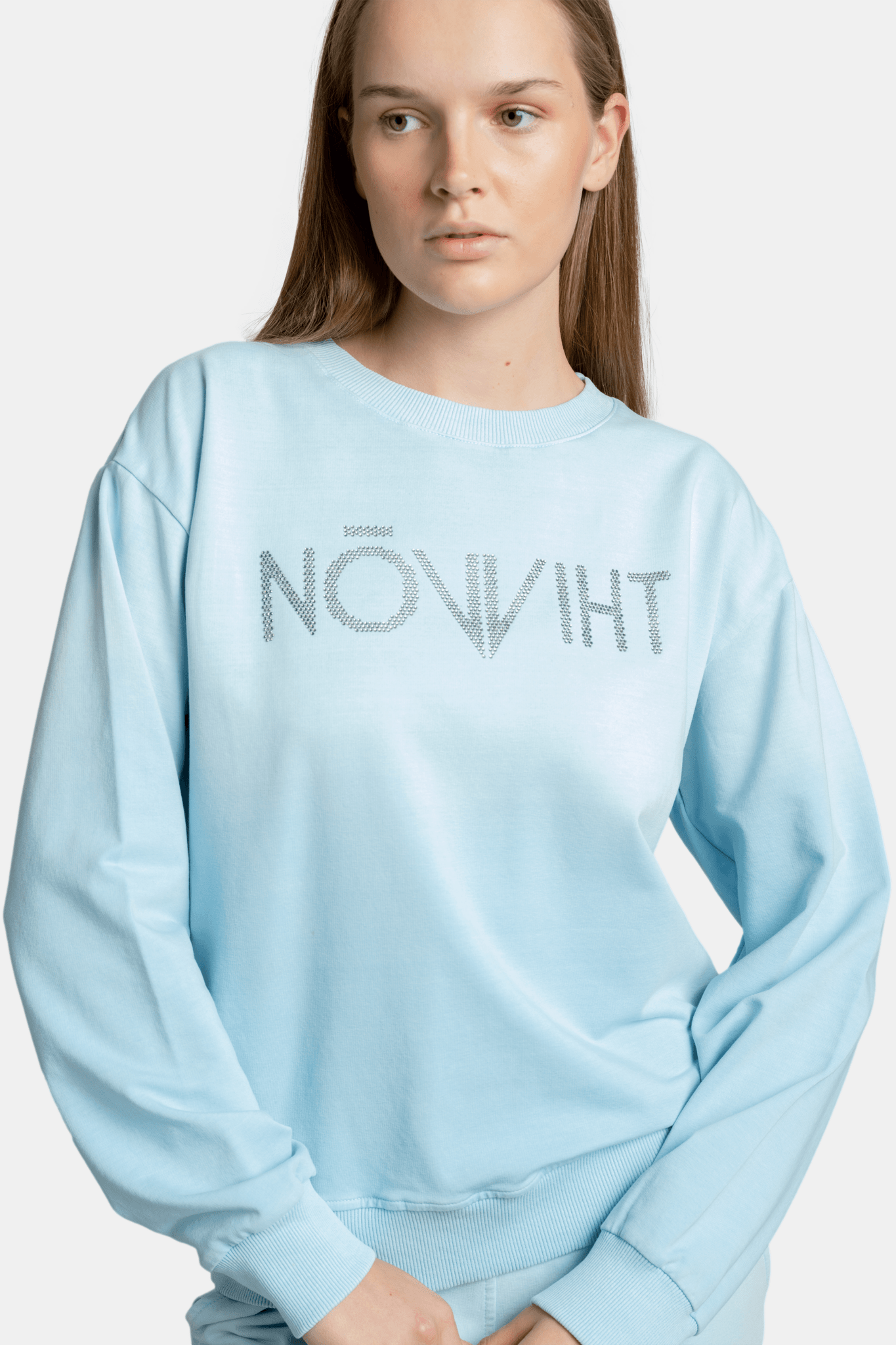 Woman in a light blue NOWIHT sweatshirt and matching joggers, posing with a relaxed posture and hands in pockets.