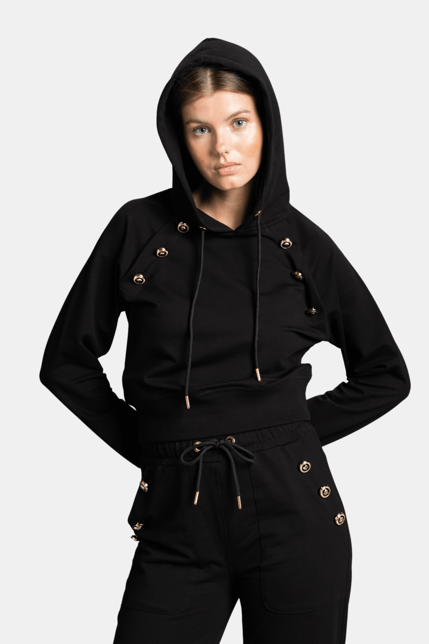 Woman in a black hoodie and pants set with gold button details, looking confidently at the camera, hood up.